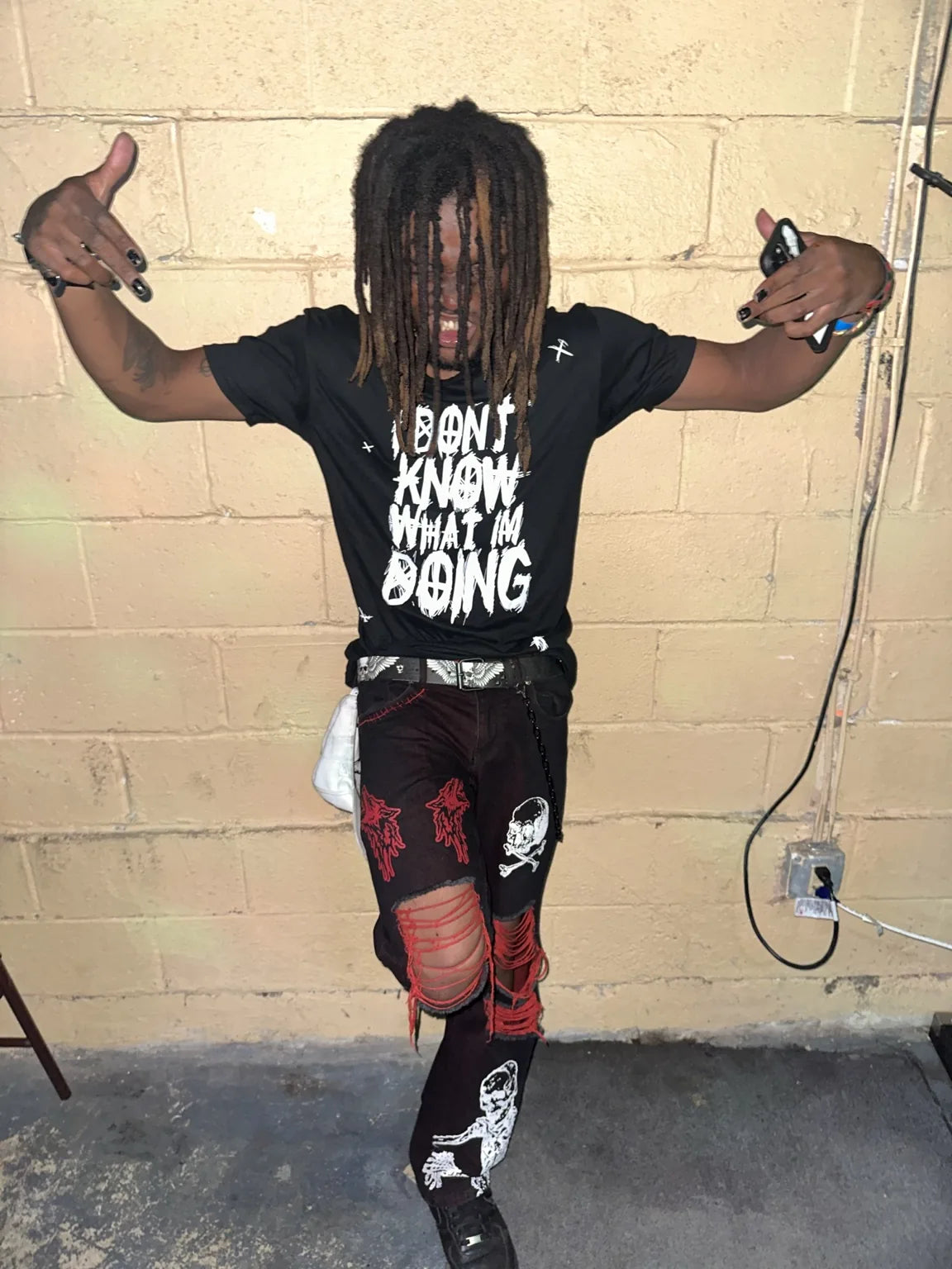 A person wearing a black Rizo graphic T-shirt against a worn block wall