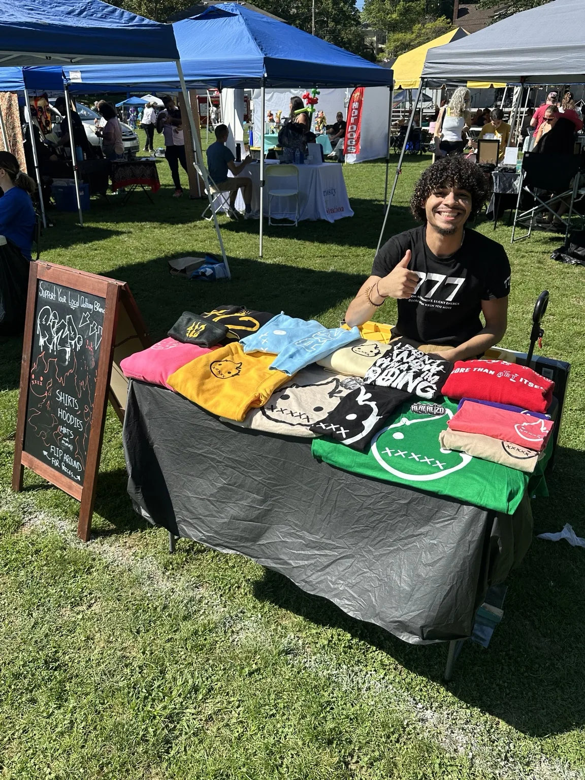 A person seated behind a Rizo pop-up table covered with colorful shirts and hoodies at an outdoor event