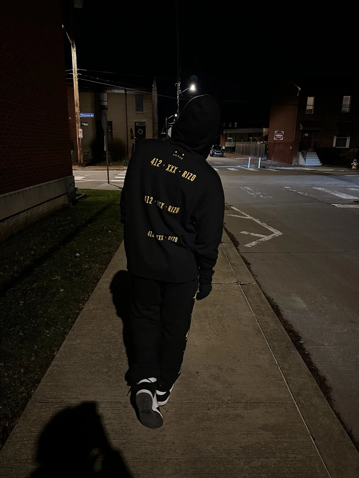 A person walking down a street at night wearing a black hoodie with yellow 412, XXX and Rizo lettering on the back