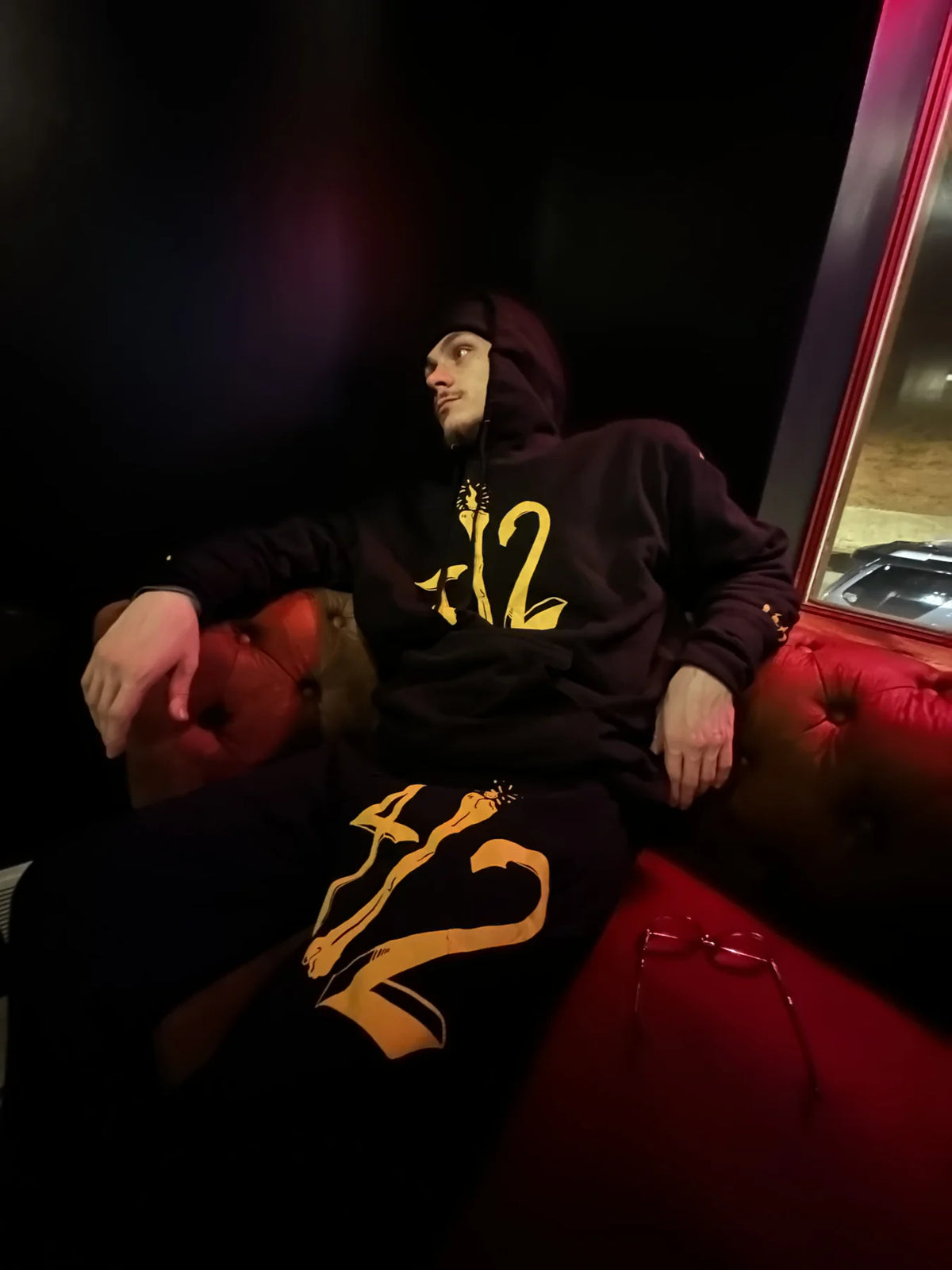 Knoah wearing a black and yellow Rizo 412 hoodie and sweats while seated on a red leather couch