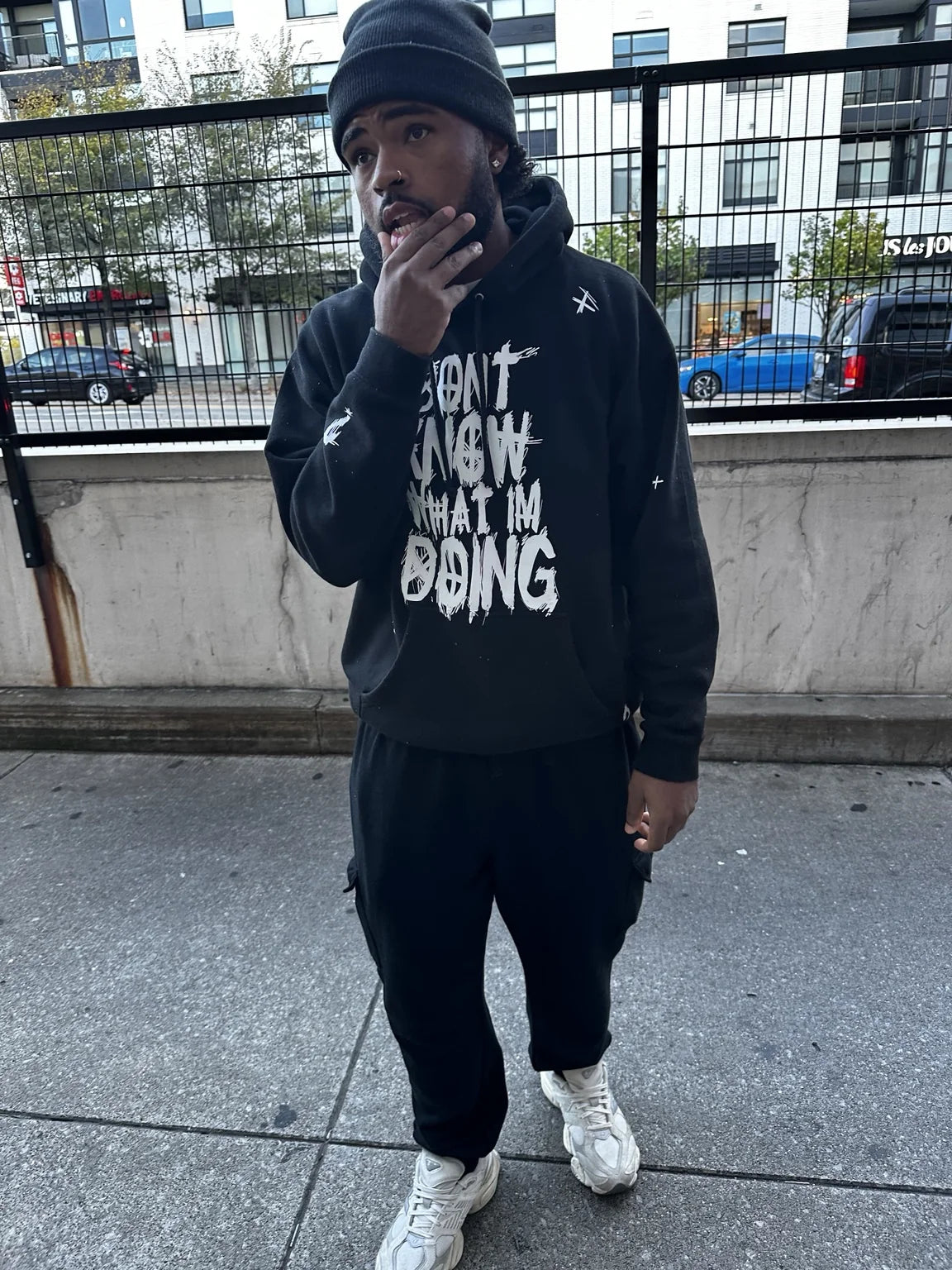 A person wearing a black Rizo graphic hoodie and knit hat on a city sidewalk