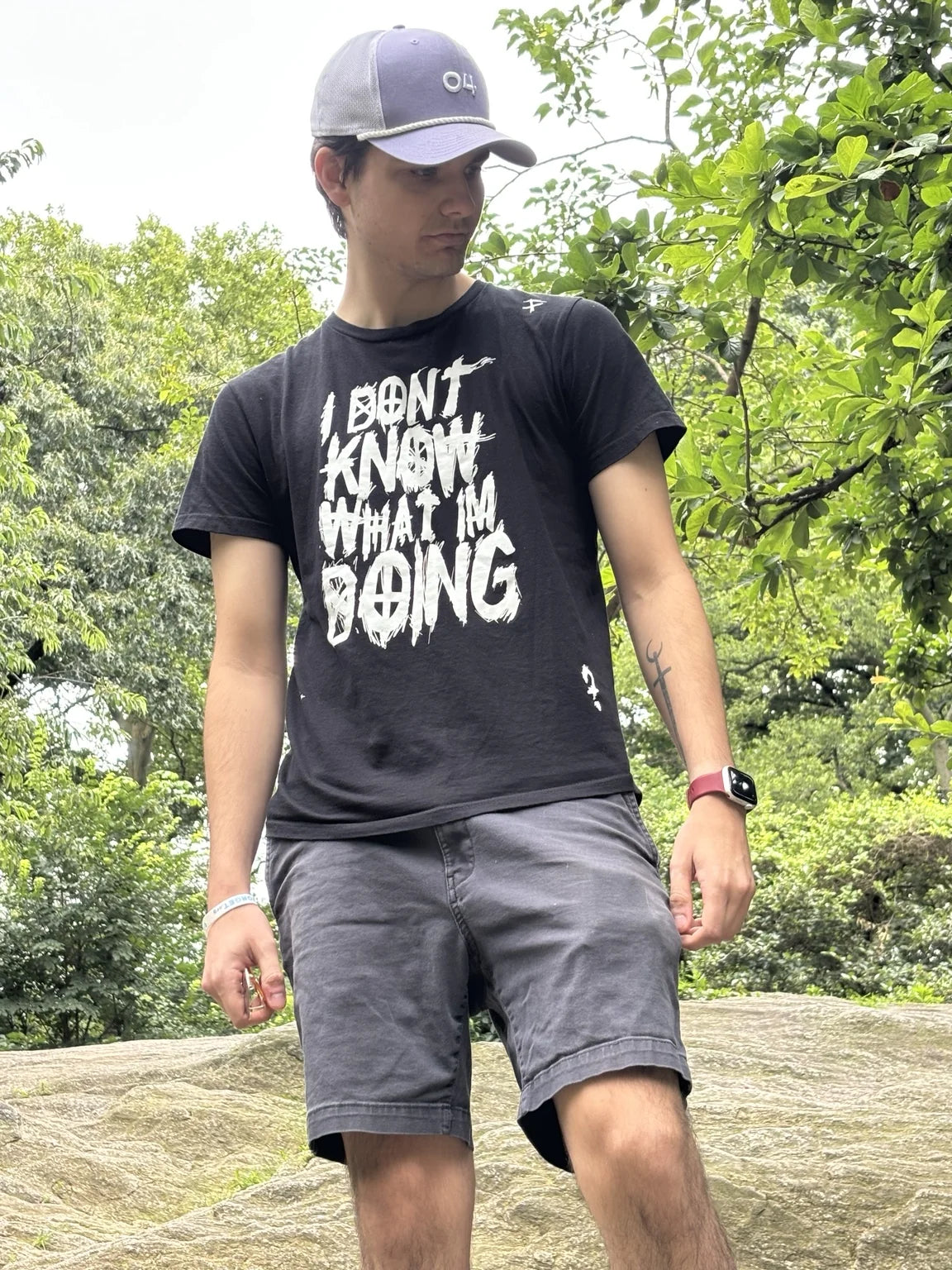 A person wearing a cap and black Rizo graphic T-shirt beside trees outdoors