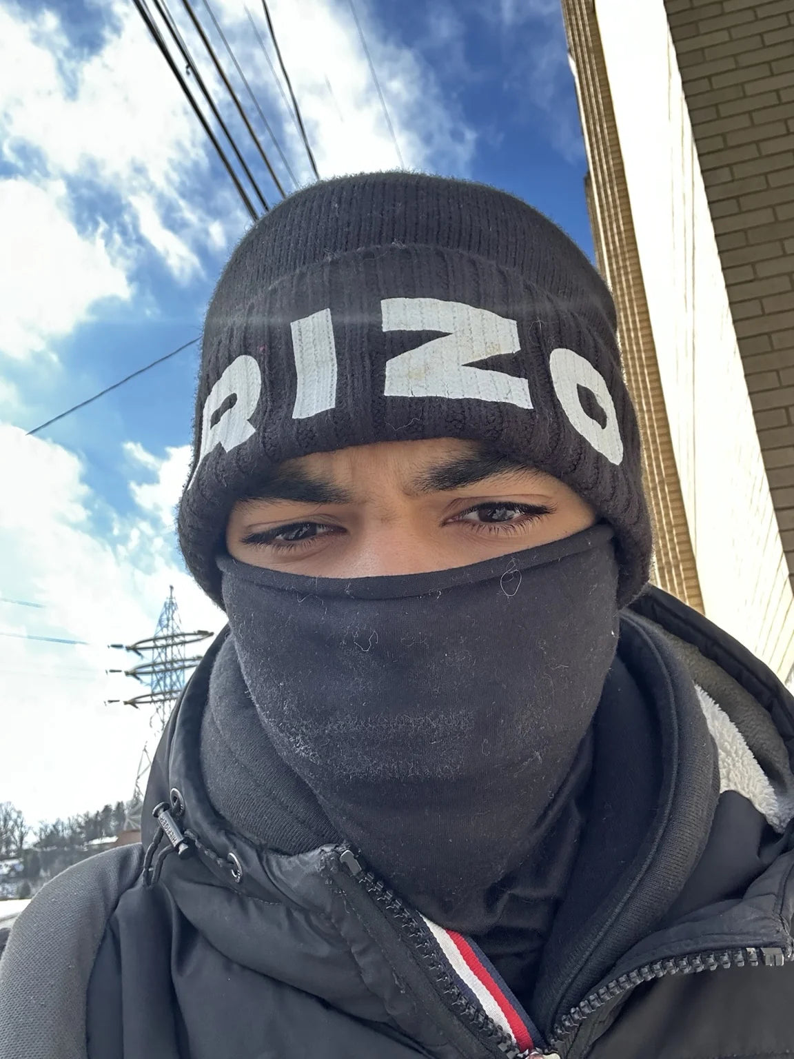 A close-up of a person outdoors wearing a black knit hat with white Rizo lettering and a face covering