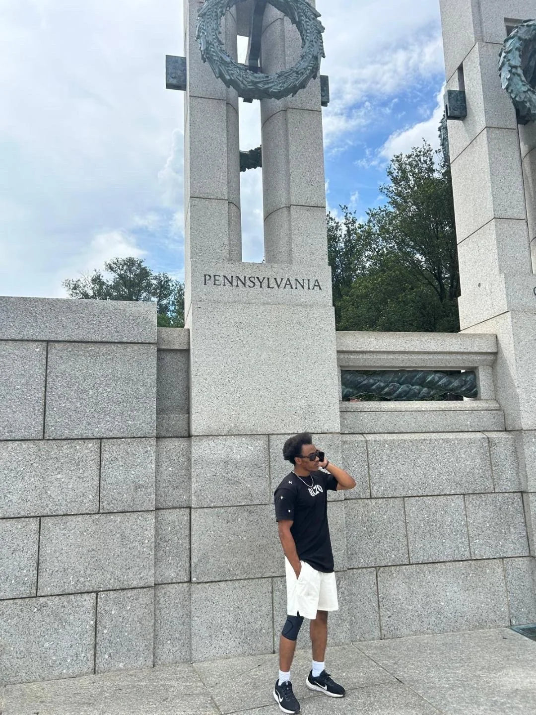 A person wearing a black Rizo T-shirt beside a stone monument with Pennsylvania lettering