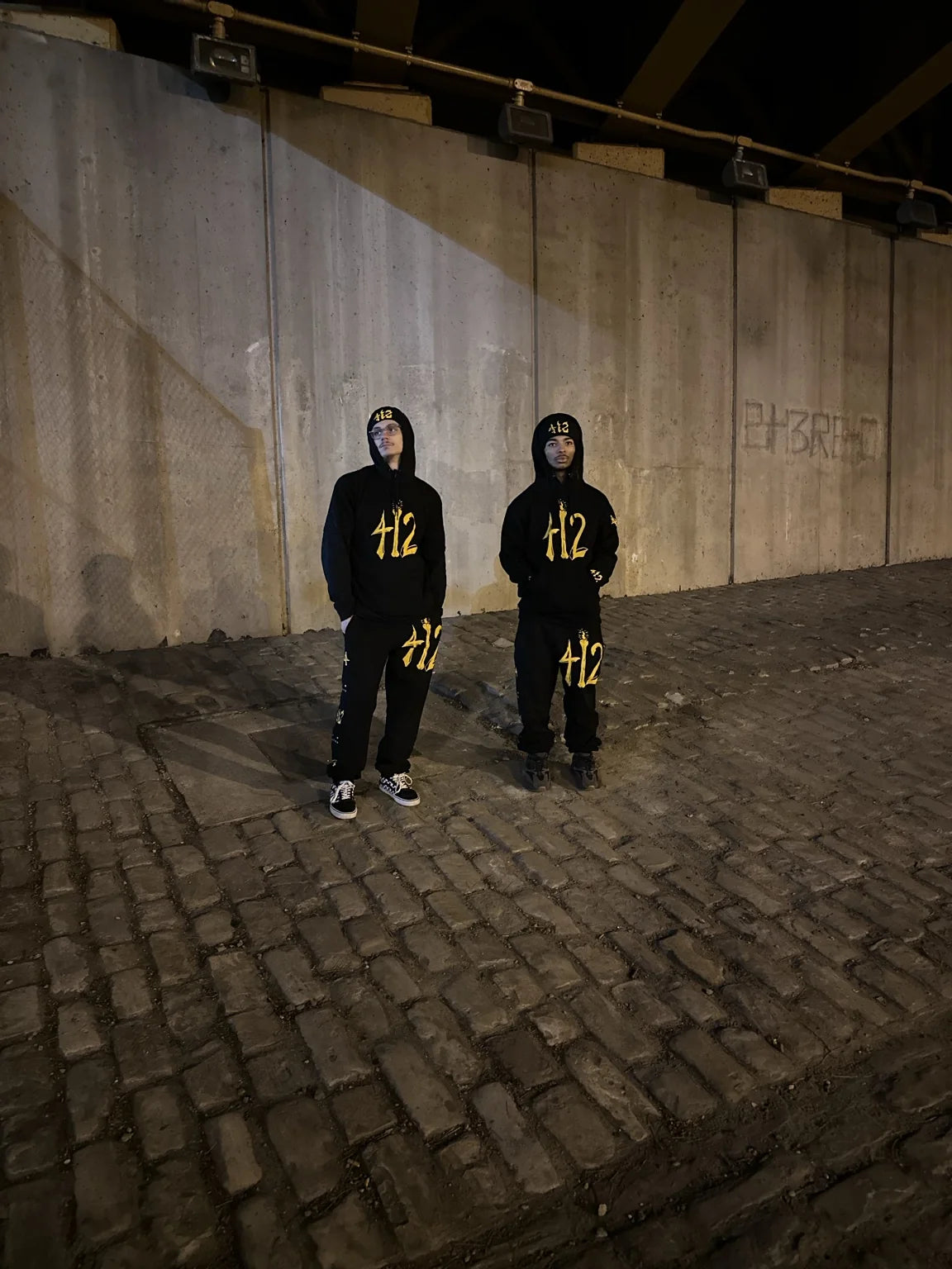 Two people wearing coordinated black and gold Rizo 412 sets beneath a Pittsburgh underpass