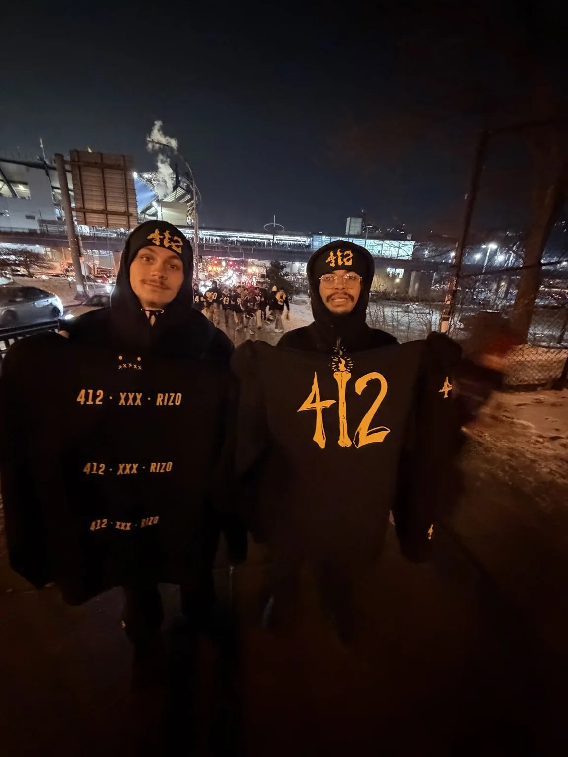 Two people wearing black and yellow Rizo 412 pieces near a Pittsburgh stadium at night